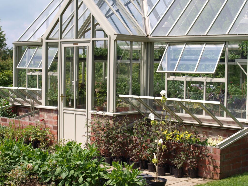 Glass greenhouse with brick base, open vent windows, and a variety of potted plants and flowers growing inside and around the structure.