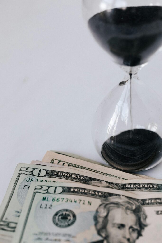 Hourglass with black sand next to US twenty-dollar bills, symbolizing time and money.
