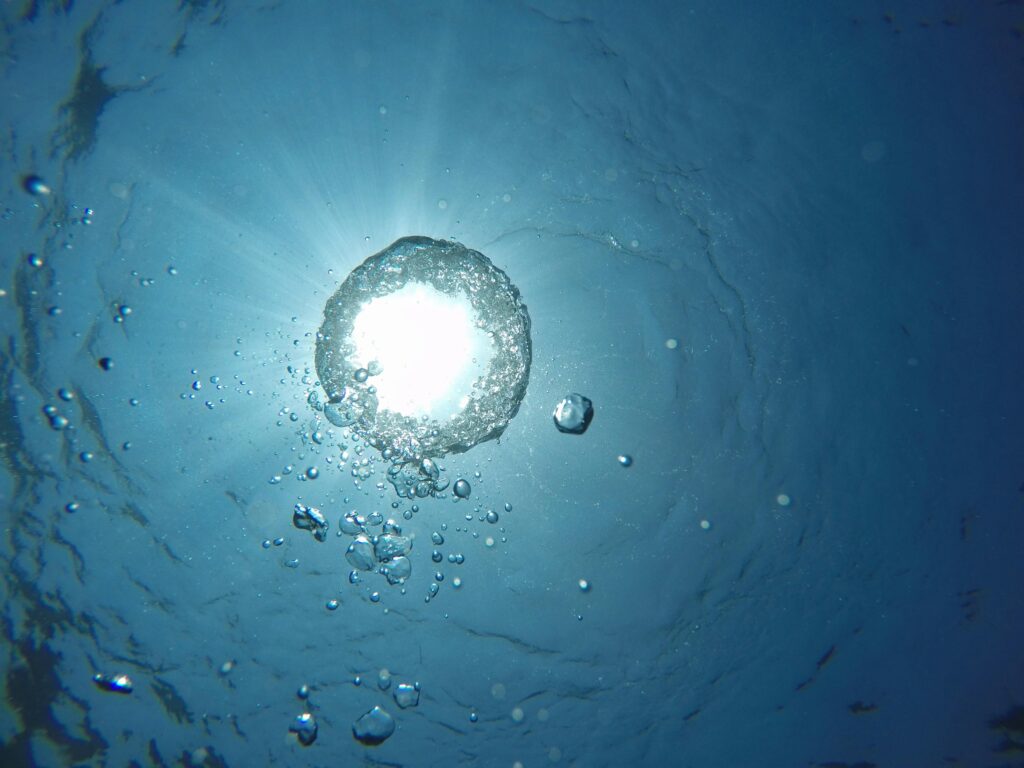 Underwater view of sunlight shining through the surface, with air bubbles rising toward the light in blue water.
