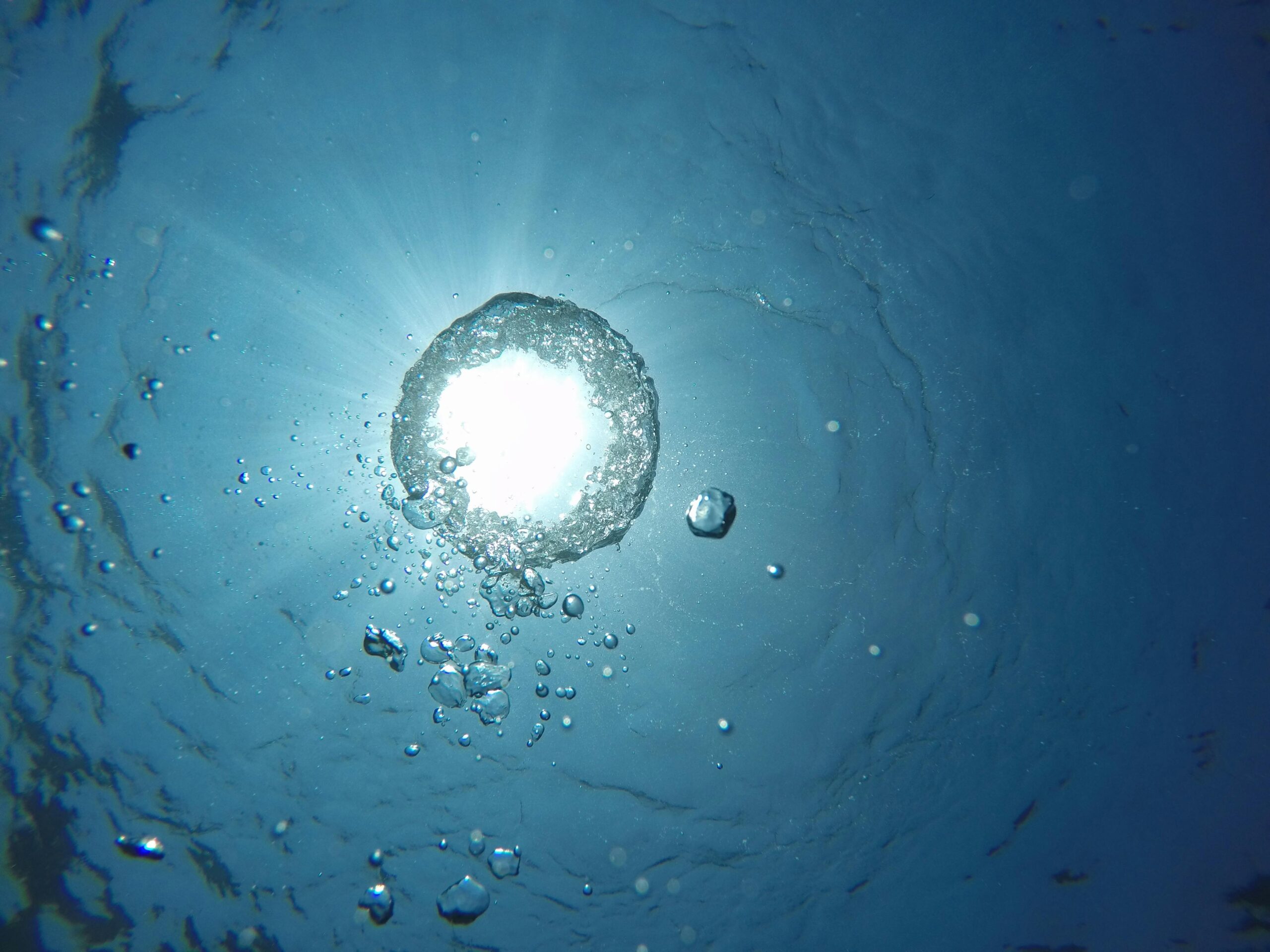 Underwater view of sunlight shining through the surface, with air bubbles rising toward the light in blue water.