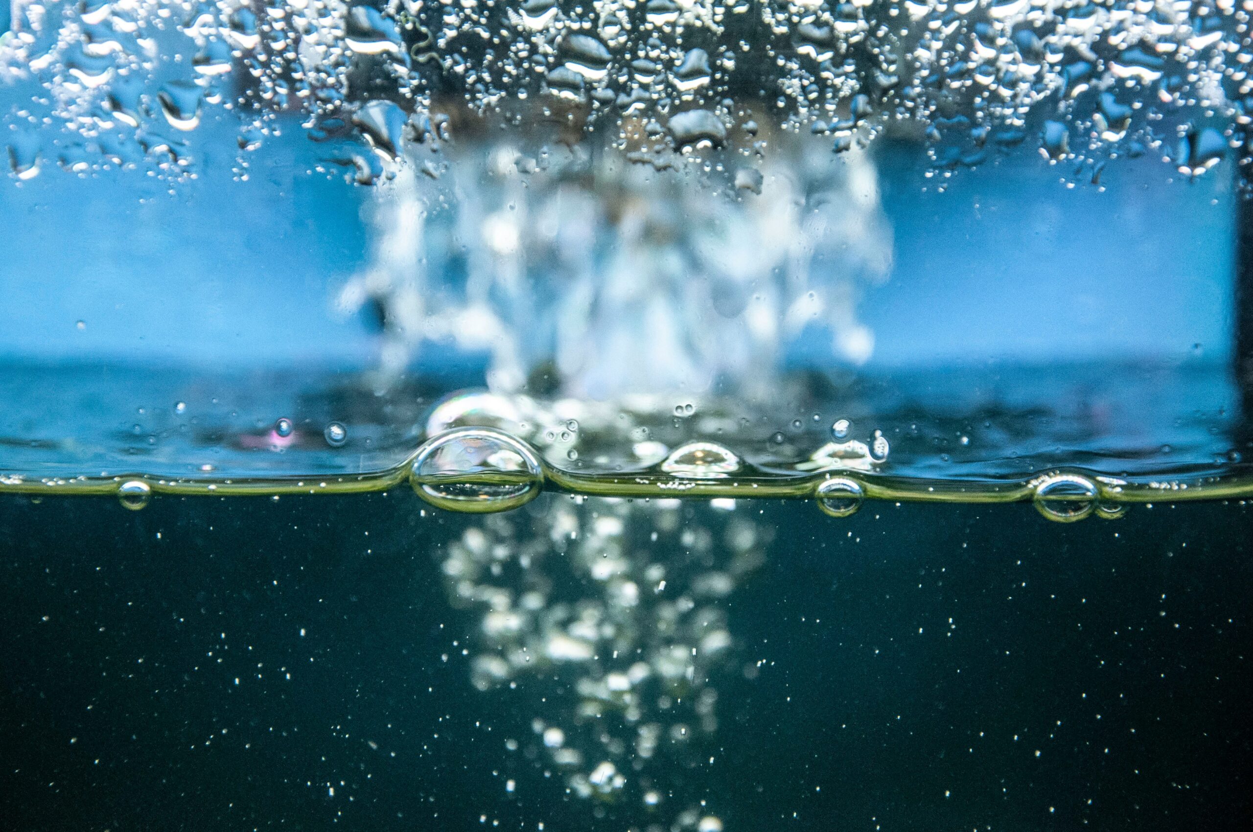 Water surface with rising air bubbles captured in a split above-and-below view, showing droplets and turbulence at the surface and a trail of bubbles dispersing underwater against a blue background.