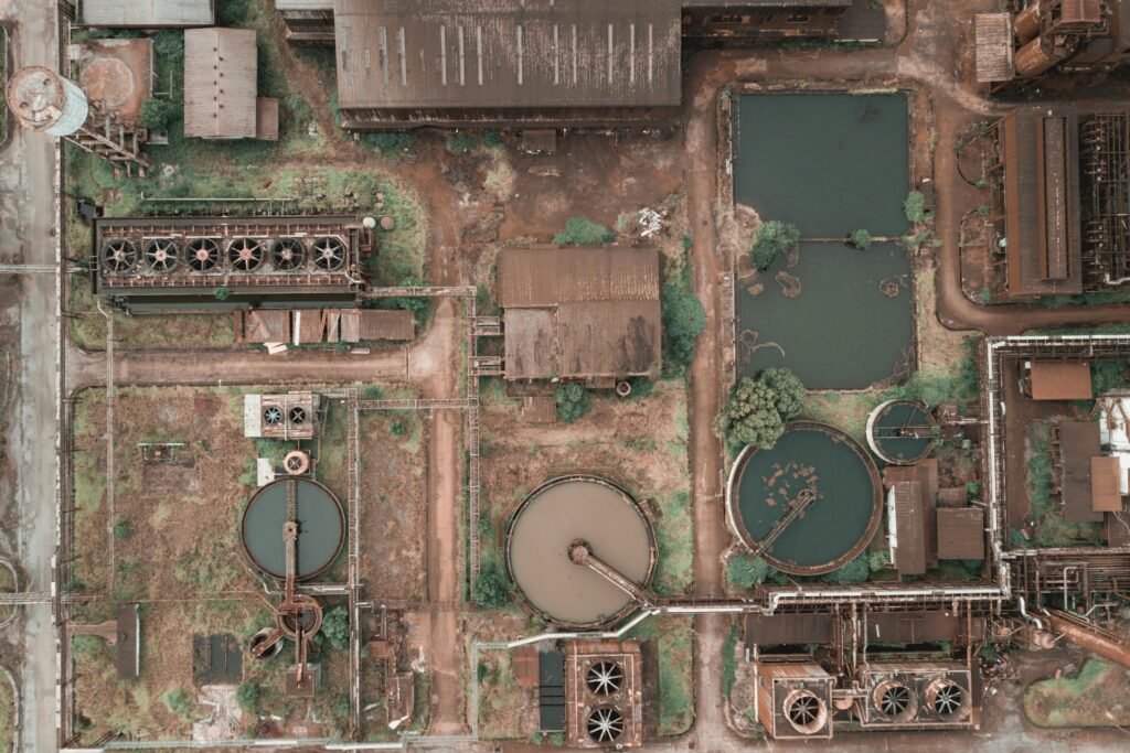 Aerial view of an industrial wastewater treatment facility with circular settling tanks, cooling systems, water basins, pipelines, and weathered factory buildings surrounded by dirt roads and vegetation.