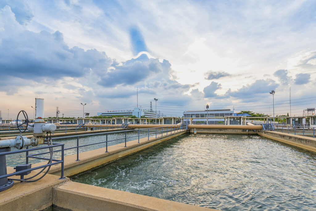 Wastewater treatment facility with open water tanks, railings, and industrial piping under a cloudy sky.