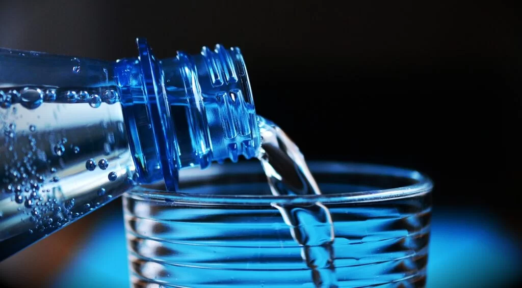 Clear water being poured from a blue plastic bottle into a drinking glass.