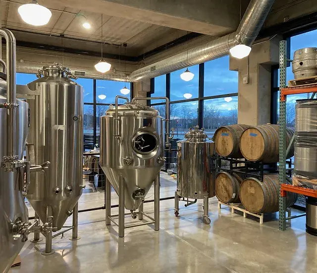 Brewery interior with stainless steel fermentation tanks, wooden barrels, and industrial brewing equipment.