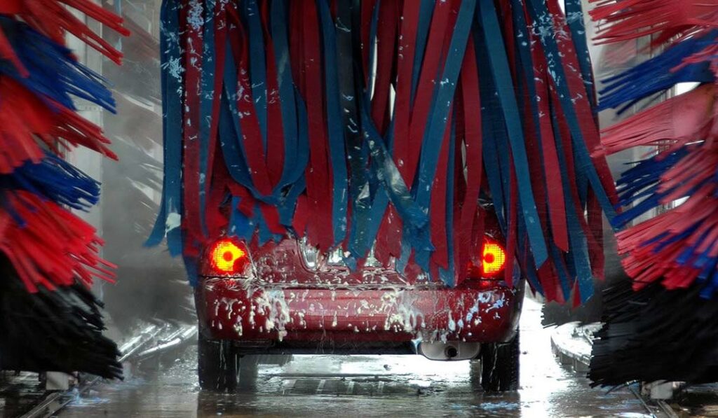 Red car moving through an automatic car wash with red and blue brushes and soap on the rear window.