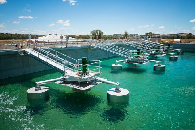 Wastewater treatment tanks with green water, floating aerators, railings, and walkways under a clear blue sky.